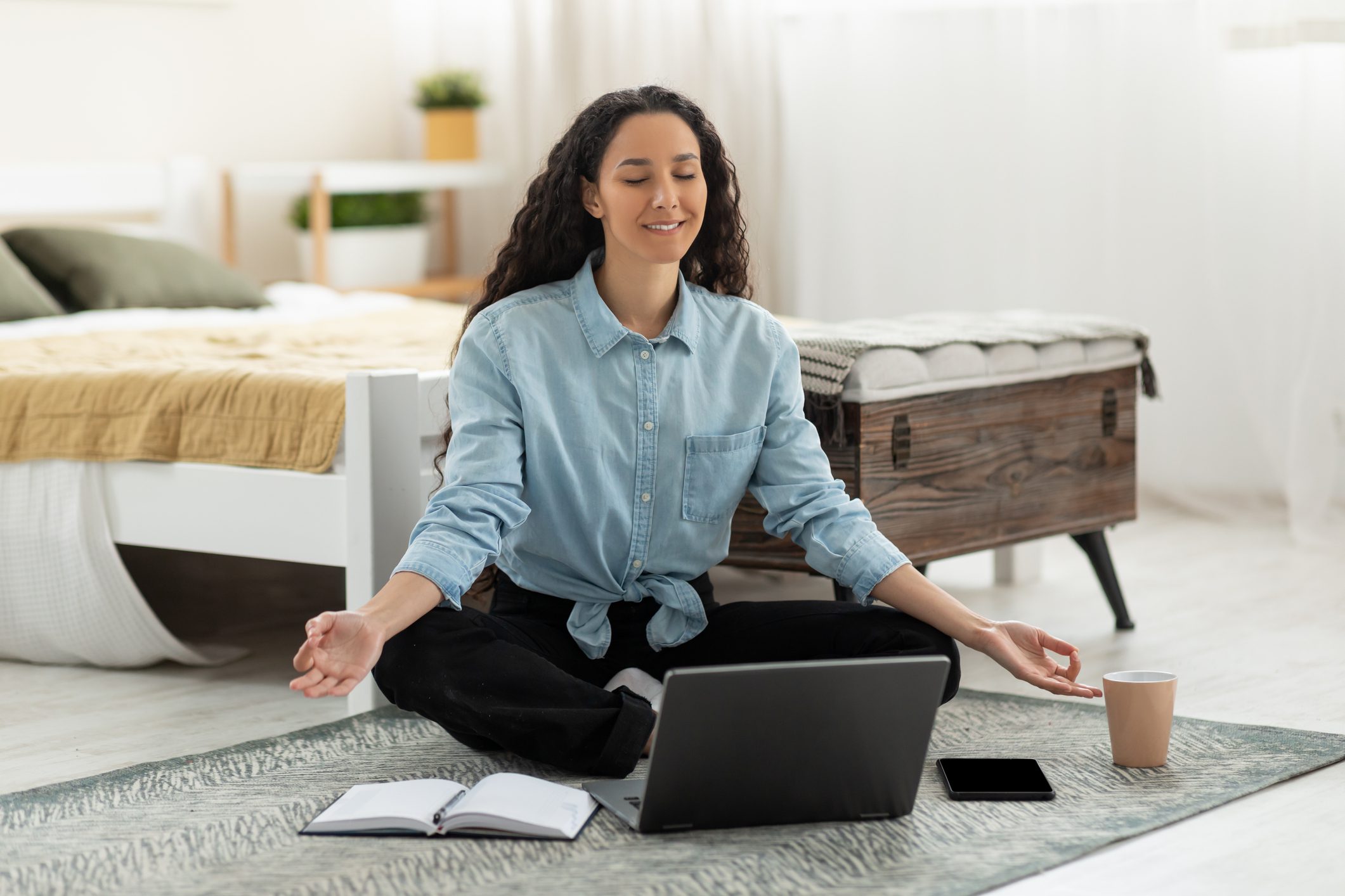 A Woman Sitting on the Floor in a Bedroom With a Laptop and a Coffee Cup, Meditating