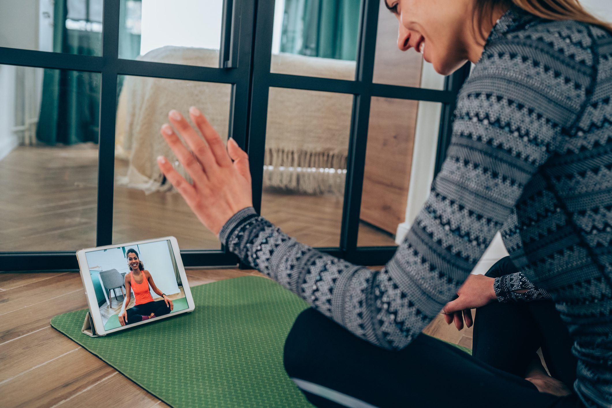 A Woman Is Sitting on a Green Yoga Mat and Waving at a Tablet Screen With a Video Call On It